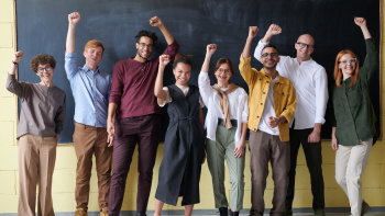 teachers of all genders and countries and skin colours lifting their arms up and smiling in front of the blackboard
