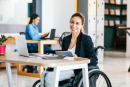 young woman in business attire on a wheelchair, working at her desk in the office and smiling towards the camera