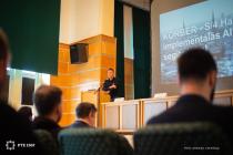 A speaker presents at a lectern in the Vargha Damján Conference Hall at the University of Pécs, in front of an audience.