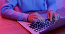Close-up of a person’s hands typing on a laptop keyboard, lit in pink and blue tones.