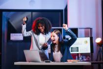 Two young people at a desk in front of a laptop, raising their fists to celebrate a success.