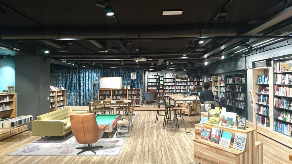 Deichman Tøyen Image of a brightly lit underground area of a library with sofas and seats in the left and center of the image and bookshelves with books classified according to genres on the right