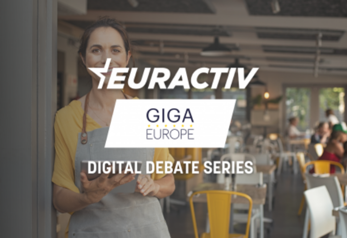A young white lady shopkeeper leaning on the door of her shop entrance looks towards the viewer, she is wearing an apron. The writing Euractiv- GIGA Europe Digital Debate Series