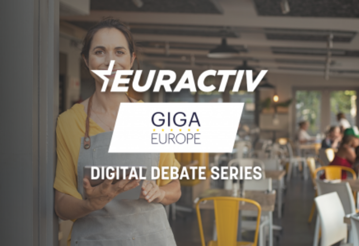 @ Euractiv A young white lady shopkeeper leaning on the door of her shop entrance looks towards the viewer, she is wearing an apron. The writing Euractiv- GIGA Europe Digital Debate Series