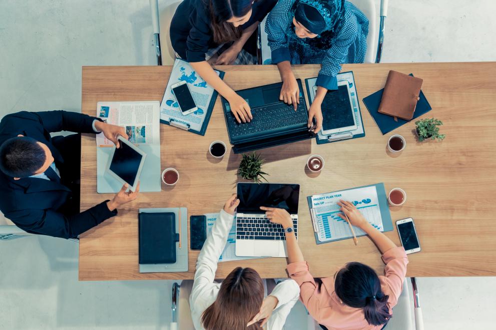 © Blue Planet Studio - stock.adobe.com Top view of businessman executive in group meeting with other businessmen and businesswomen in modern office with laptop computer, coffee and documents