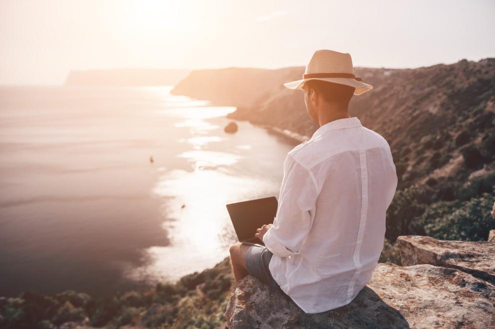 Photo of a man working on a laptop while sitting on a rock in the sunset | panophotograph Photo of a man working on a laptop while sitting on a rock in the sunset | panophotograph