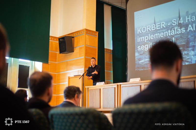 A speaker presents at a lectern in the Vargha Damján Conference Hall at the University of Pécs, in front of an audience.