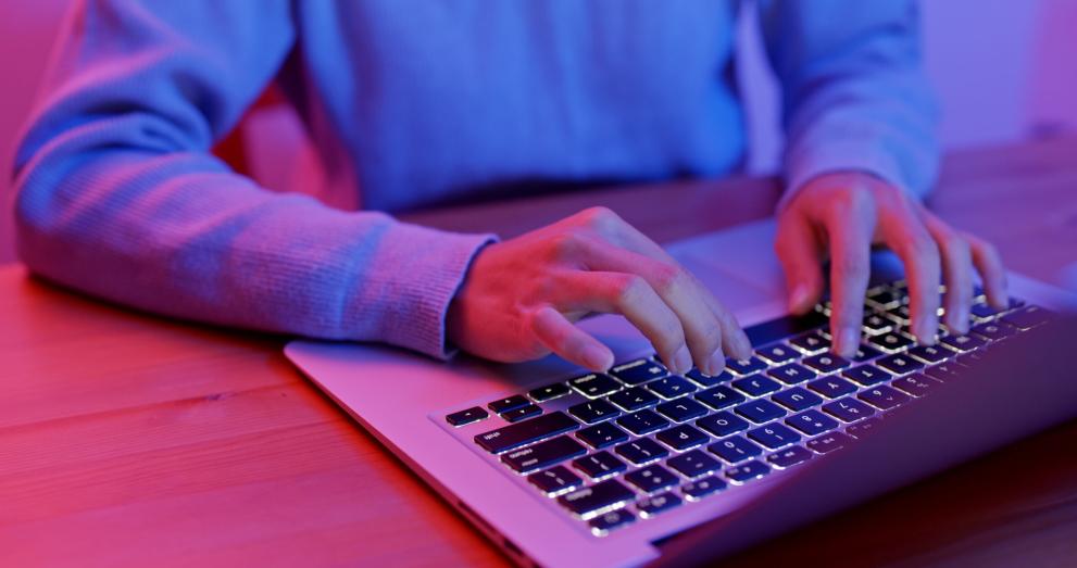 Close-up of a person’s hands typing on a laptop keyboard, lit in pink and blue tones.