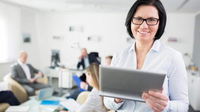 Close up photo of a middle aged business woman holding a tablet. Background is blurry with a multigenerational team sitting down around low tables with documents and computers in front of them.