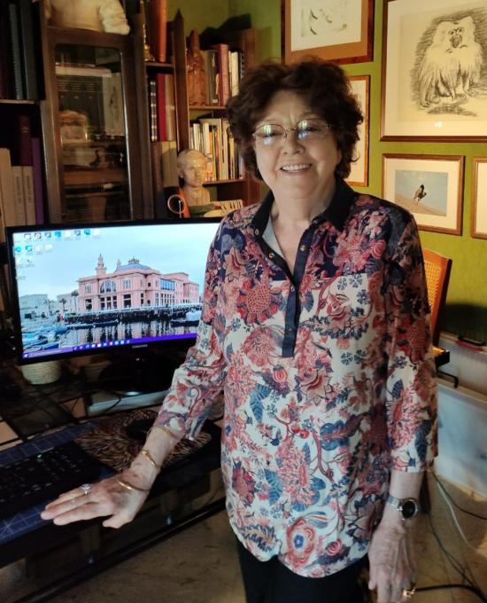 Clotilde, an Italian lady with dark hair and a colourful shirt posing in her living room in front of her computer