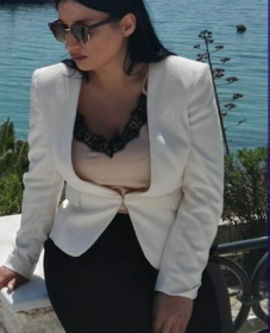 Christina, a young woman with dark long hair, sunglasses, wearing a formal jacket and trousers and posing in front of a beautiful blue sea