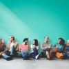 A group of people sitting against a wall, with laptops and devices collaborating. 
