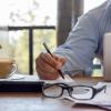A man writing on a notebook with a cup of coffee and his glasses by his side. 