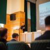 A speaker presents at a lectern in the Vargha Damján Conference Hall at the University of Pécs, in front of an audience.