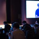 Presenter with Hand Up Giving Presentation while Audience People Watch in Conference Hall Auditorium. Blurred De-focused Unidentifiable Presenter and Audience.